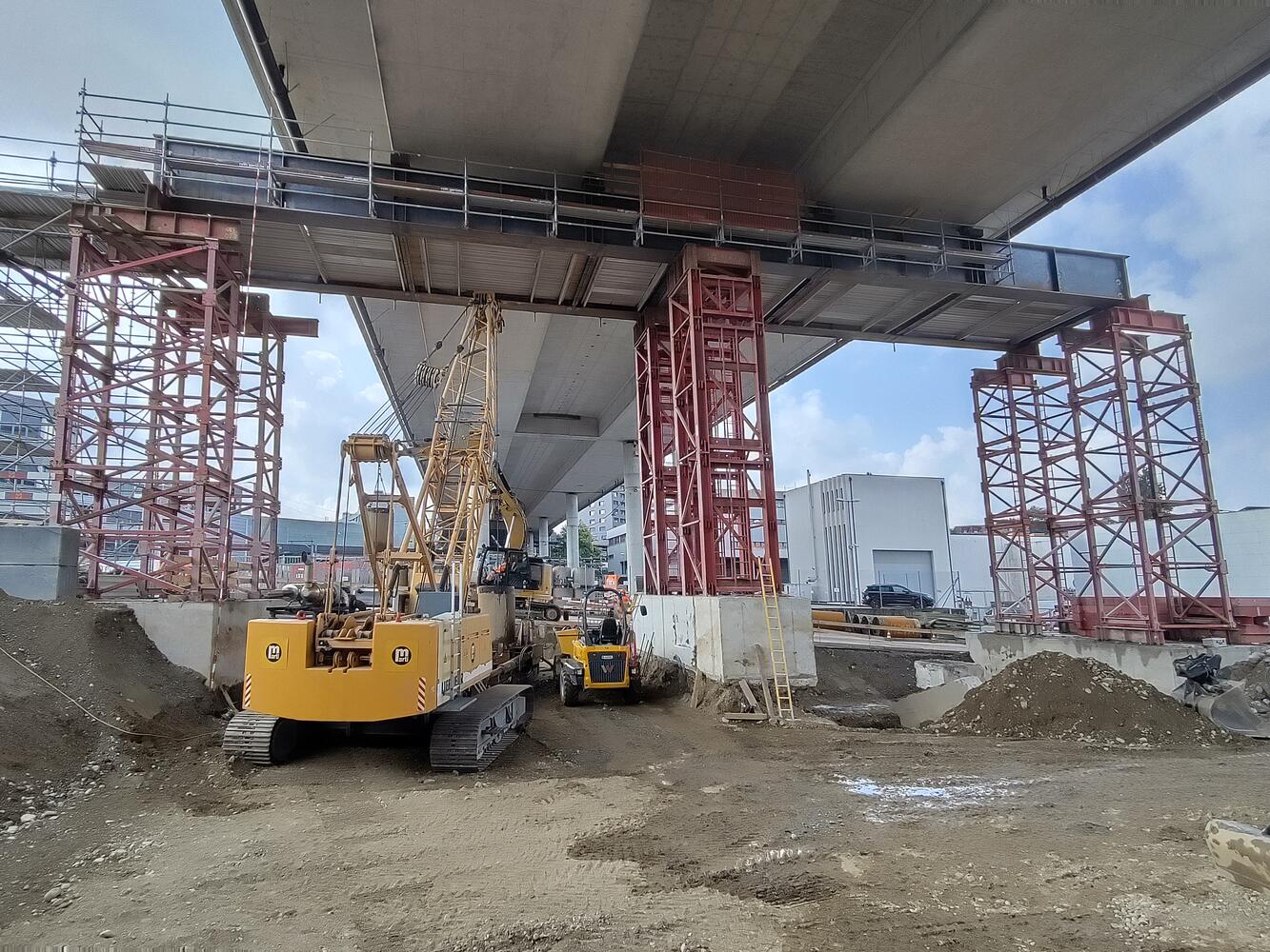 Bild einer Baustelle unter einer Brücke mit Stahlträgern und einem Bagger.