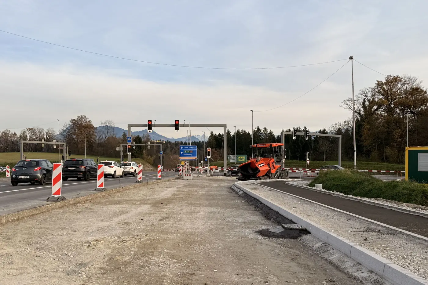 Baustelle an einer Straße mit Ampeln und Baumaschinen, Wolken am Himmel.