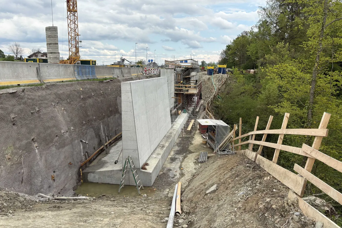 Baugrube mit Betonwand, Baustellenmaterial und Bäumen im Hintergrund.