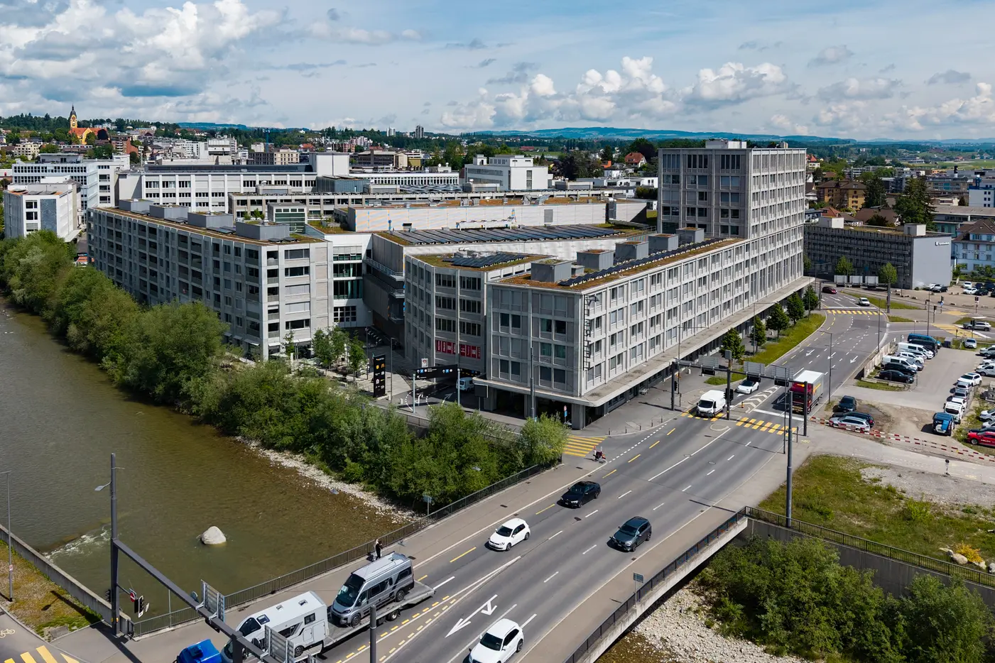 Gebäudekomplex mit Strasse auf welcher Autos ersichtlich sind, links vom Gebäude ist ein Fluss mit Bäumen zu sehen
