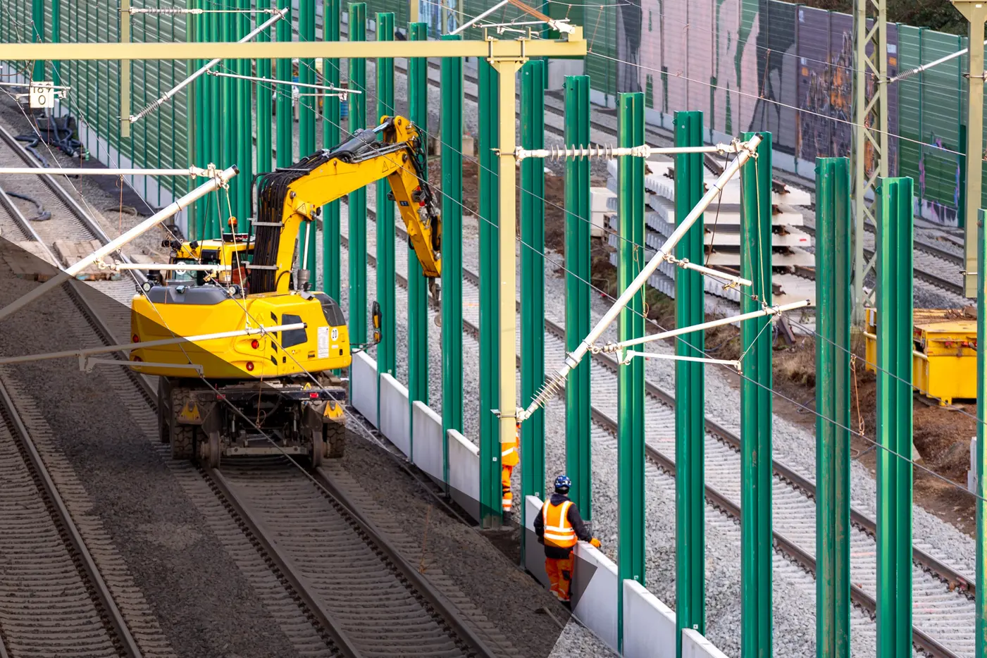 Baumaschinen und Arbeiter installieren grüne Stützpfeiler neben den Gleisen.