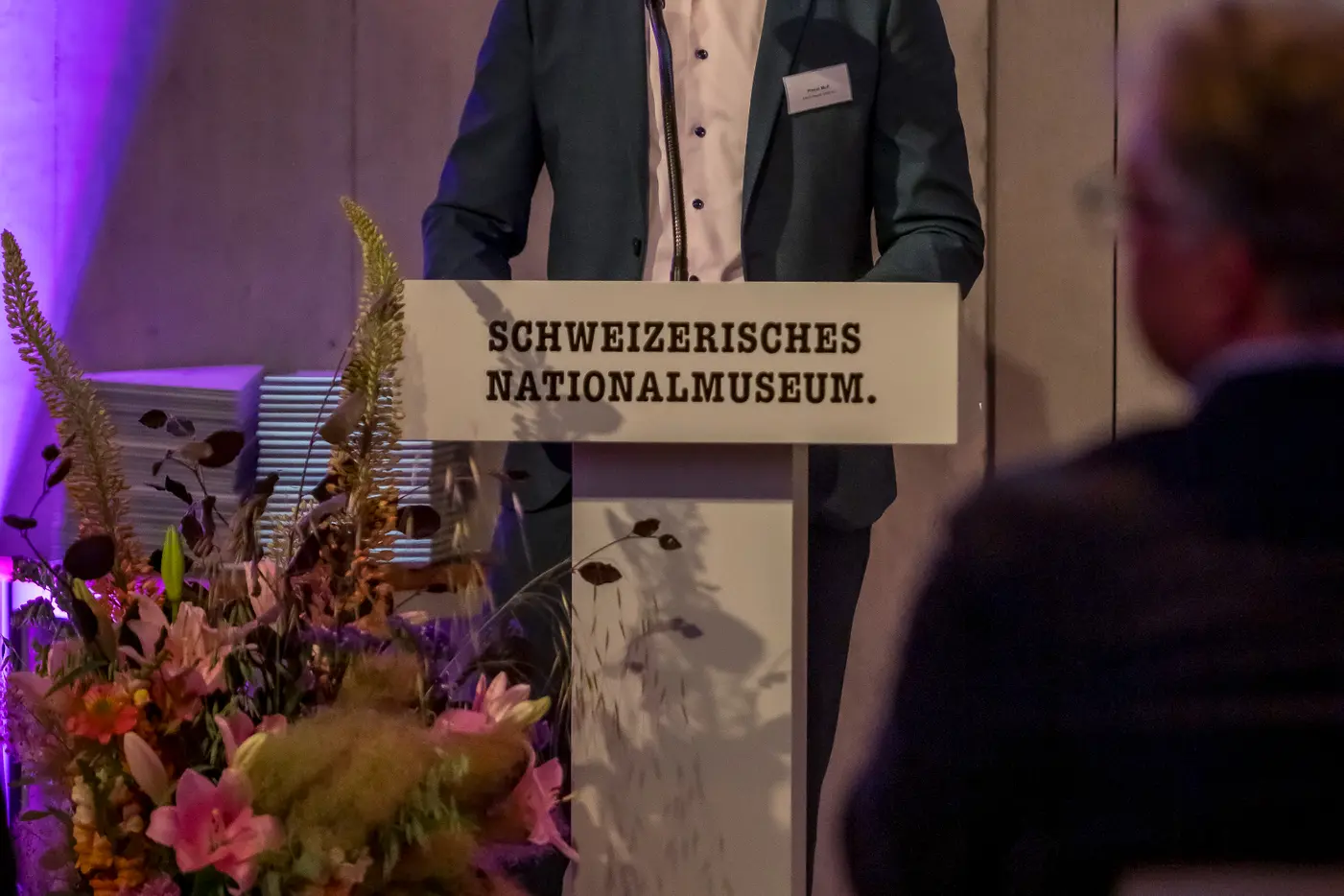 Ein redende Mann am Podium mit bunten Blumen im Hintergrund, sanfte Beleuchtung.