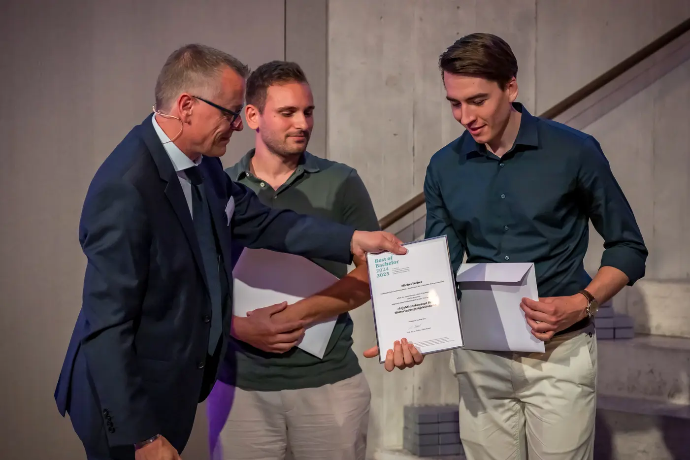 Drei Männer stehen auf einer Bühne, ein Mann überreicht einem anderen ein Zertifikat.