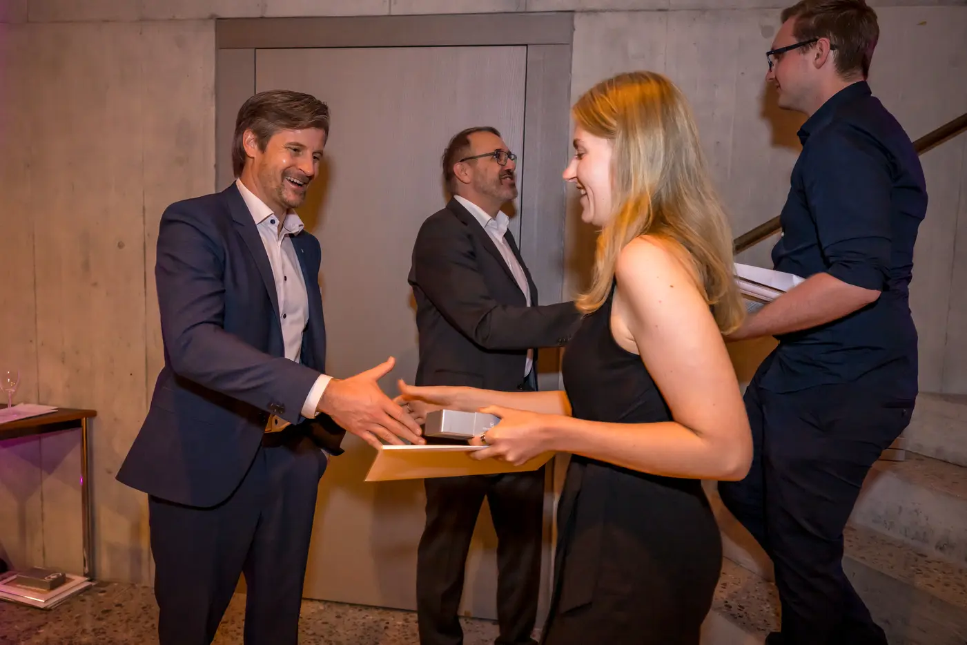 Eine Frau überreicht einem Mann ein Dokument, während zwei weitere Männer im Hintergrund stehen.