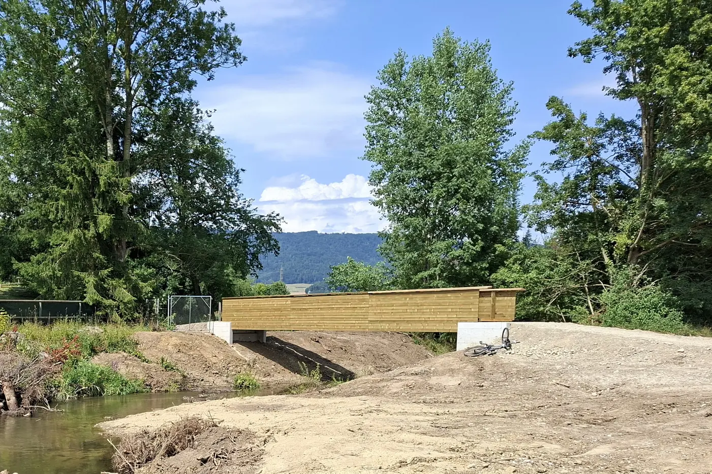 Holzbrücke über einen schmalen Fluss, umgeben von Bäumen und Bergen im Hintergrund.