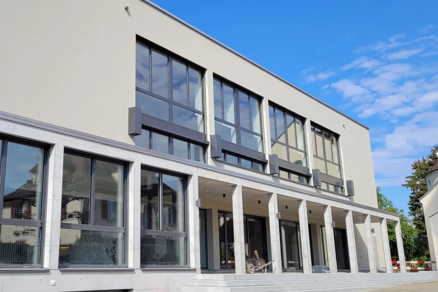 Moderne Gebäude mit großen Fenstern und überdachtem Eingangsbereich, blauer Himmel.