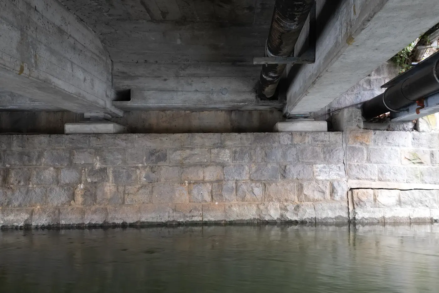 Unterseite einer Brücke mit wassernahen Steinen und Rohren.