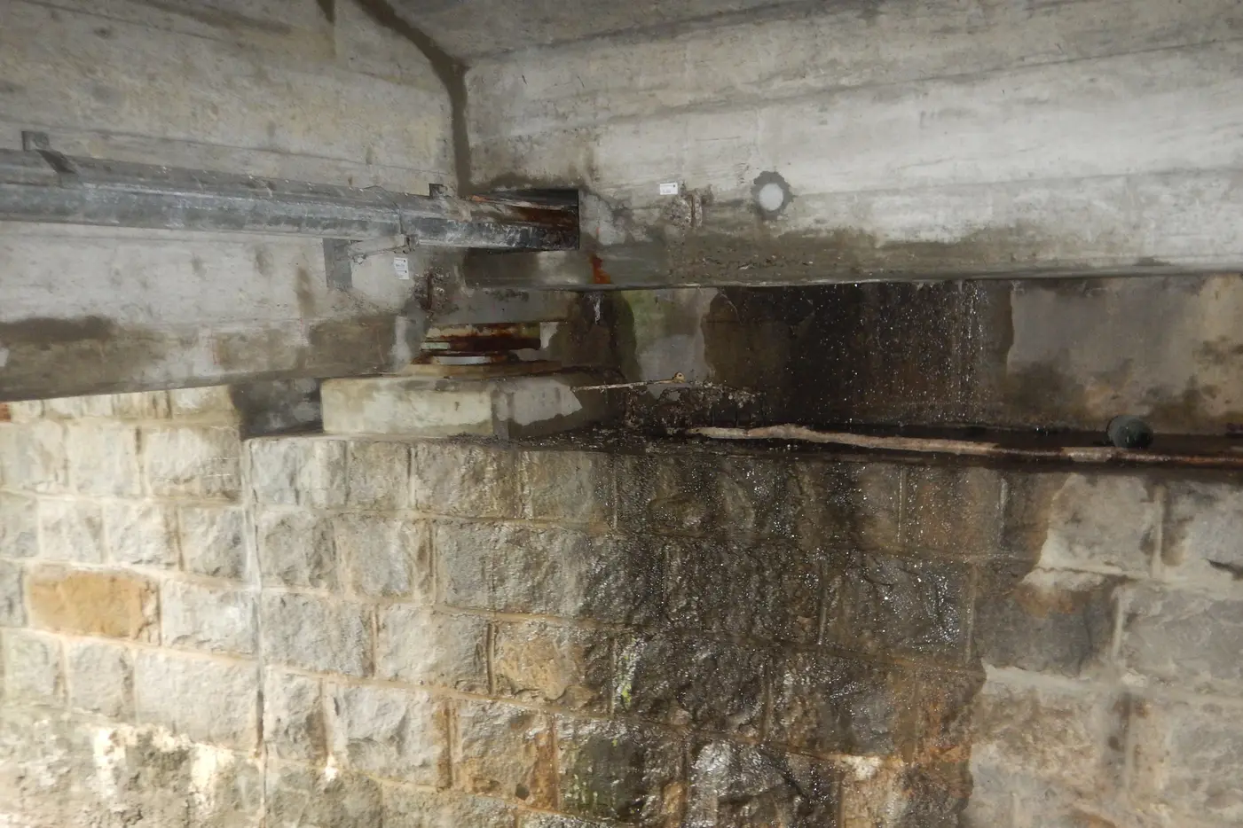 Feuchte Mauer mit sichtbaren Stahlträgern und Ziegelsteinen im Hintergrund.