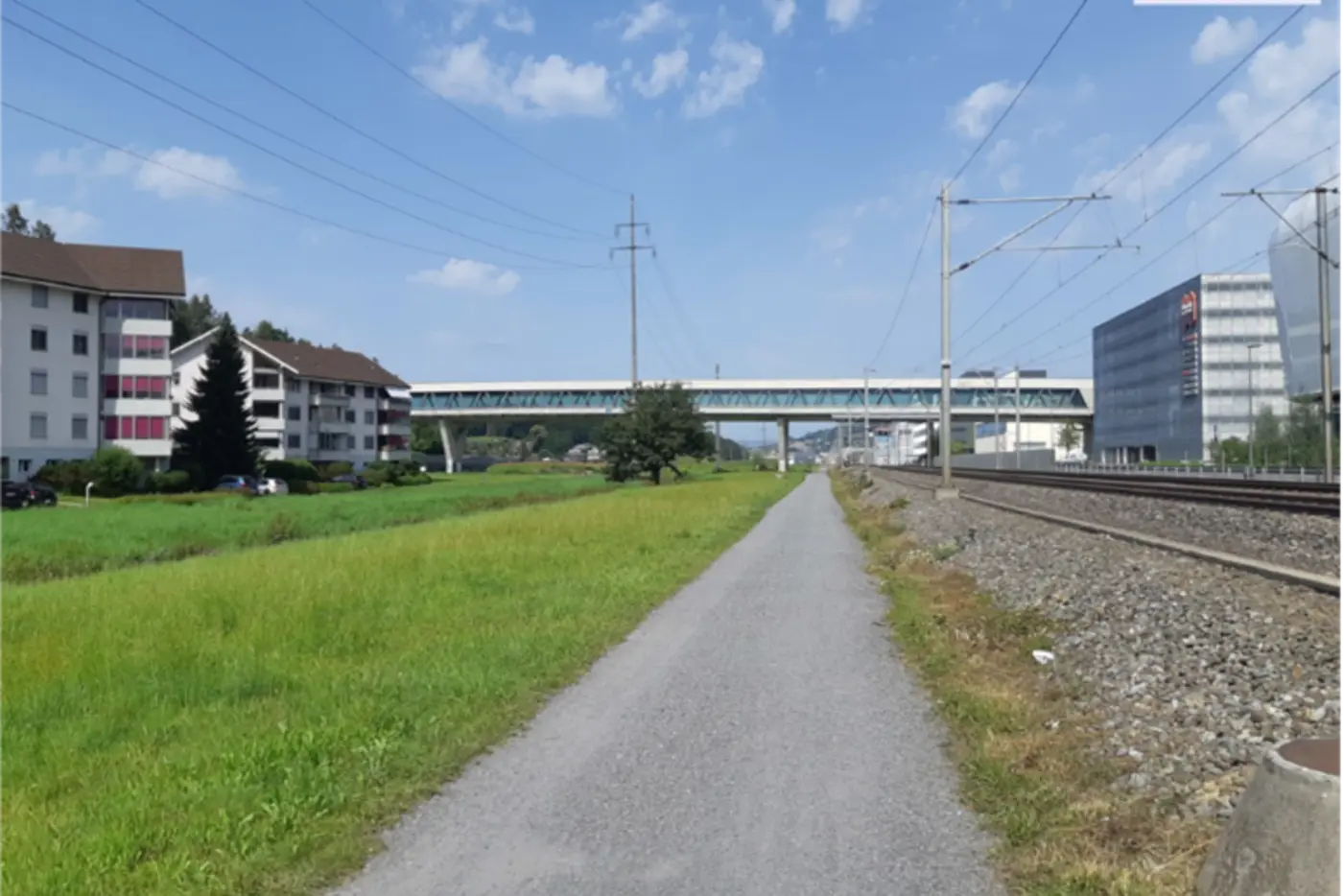Ein Radweg führt entlang von Grünflächen und Bahngleisen unter einem vierspurigen Viadukt.
