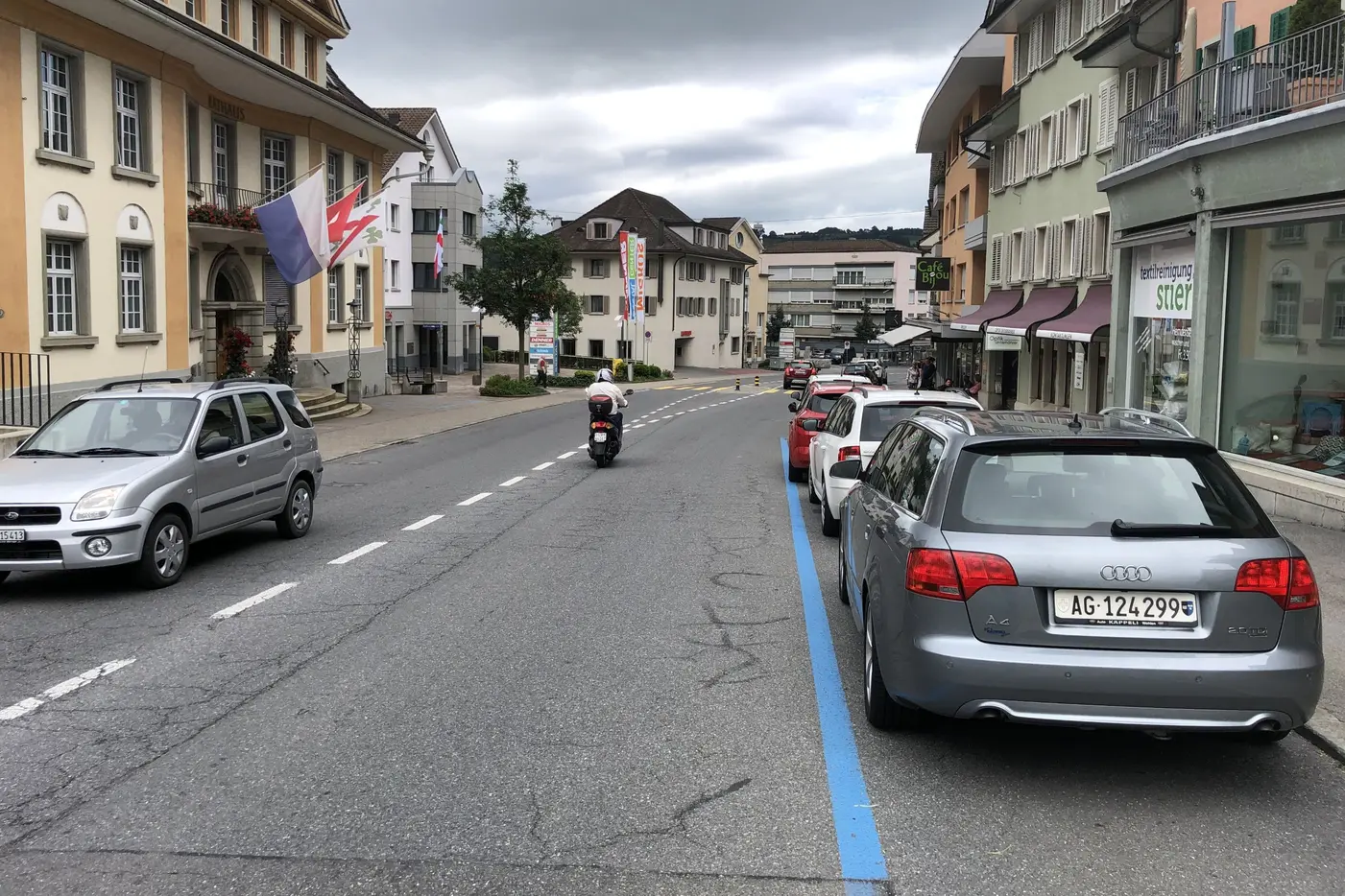 Leere Straße mit geparkten Autos und einer Motorradfahrerin unter bewölkten Himmel.