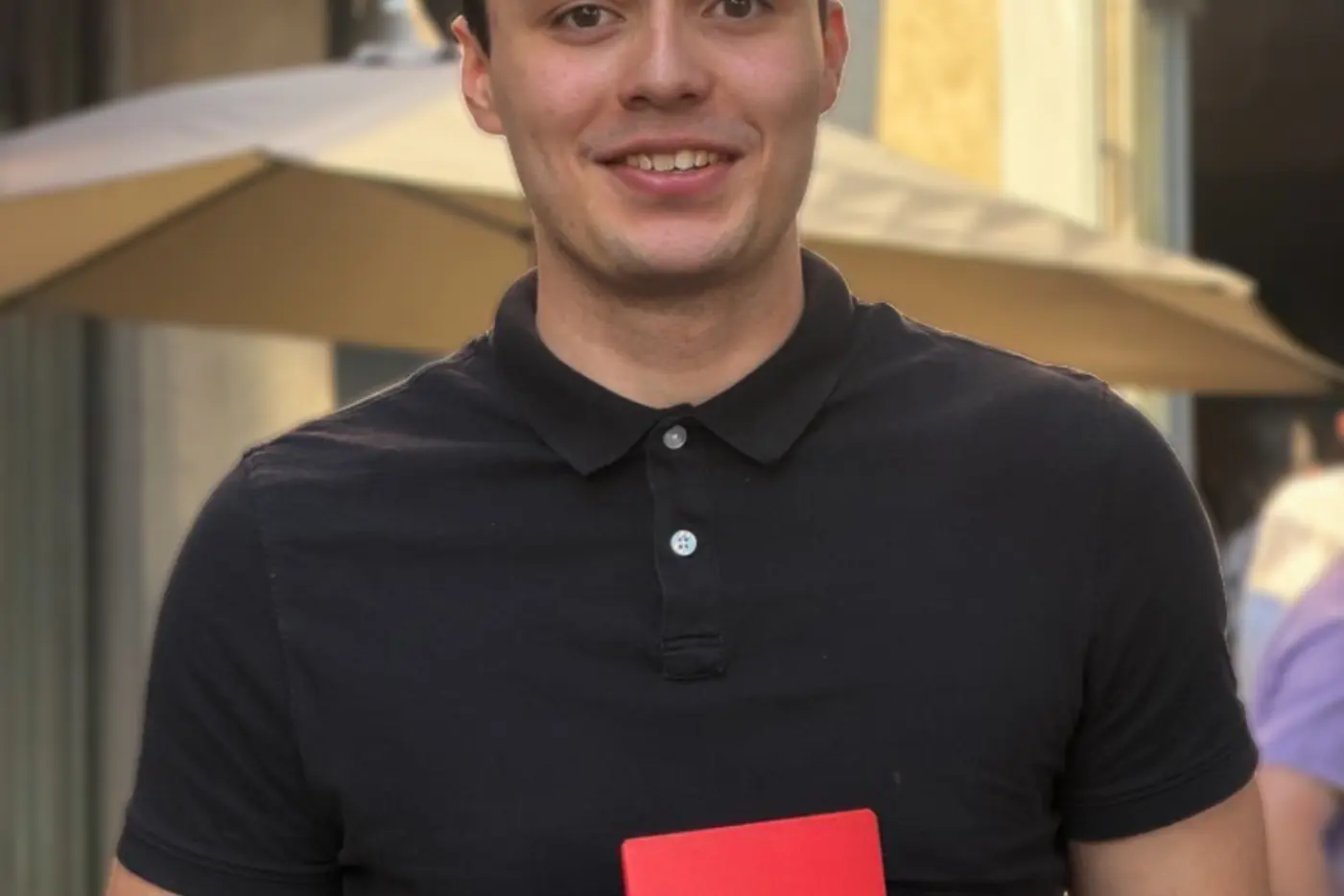 Junger Mann in einem schwarzen Polohemd hält ein rotes Heft in einem sonnigen Aussenbereich.