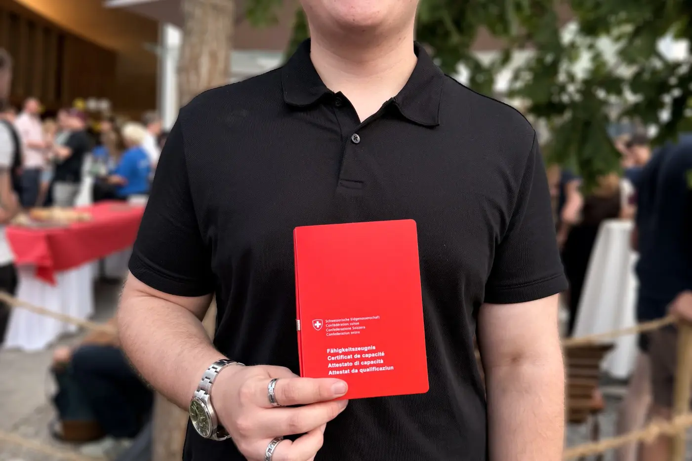 Junger Mann in schwarzem Hemd lächelt und hält ein rotes Heft in der Hand.