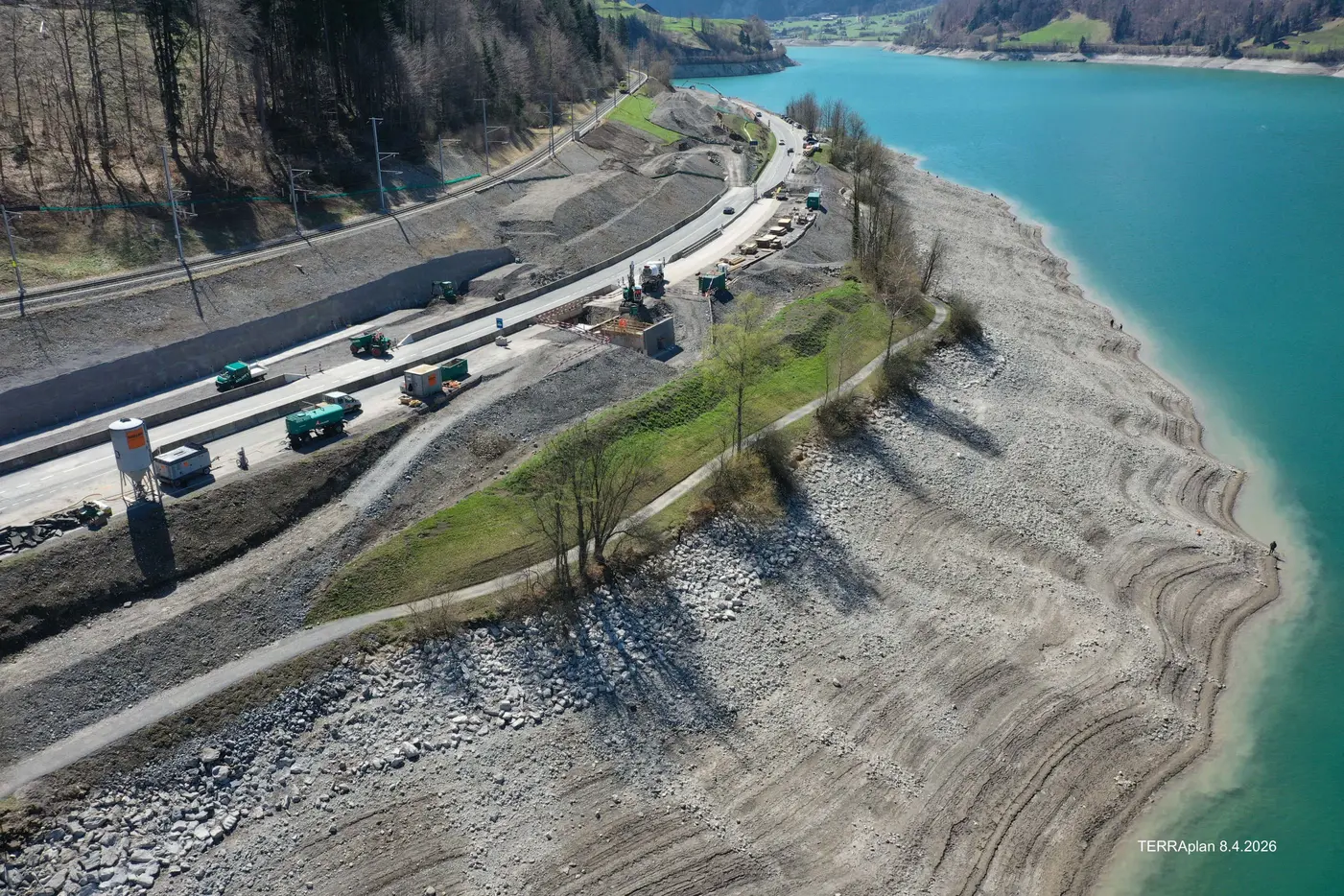 Baustelle am Ufer eines blauen Gewässers mit minimalem Wasserstand und grüner Vegetation.