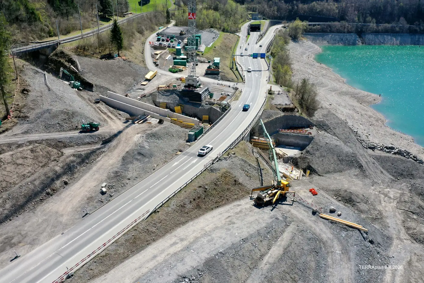 Bauarbeiten an einer Straße nahe einem Wasserlauf, graue Erde und Maschinen sichtbar.