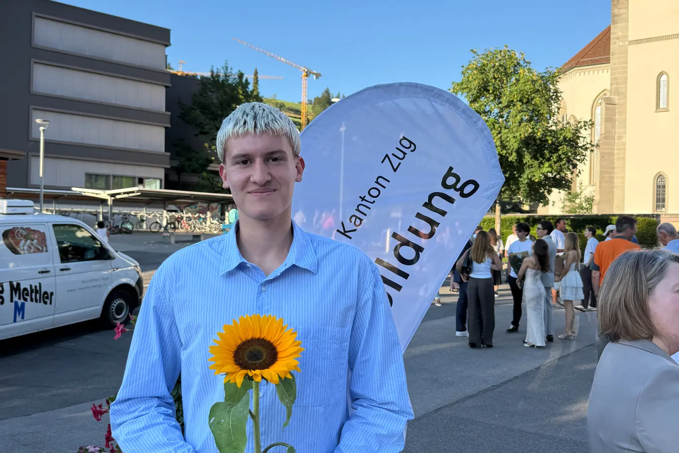 Junger Mann hält eine Sonnenblume, lächelt vor einer Menschenmenge und Gebäuden.