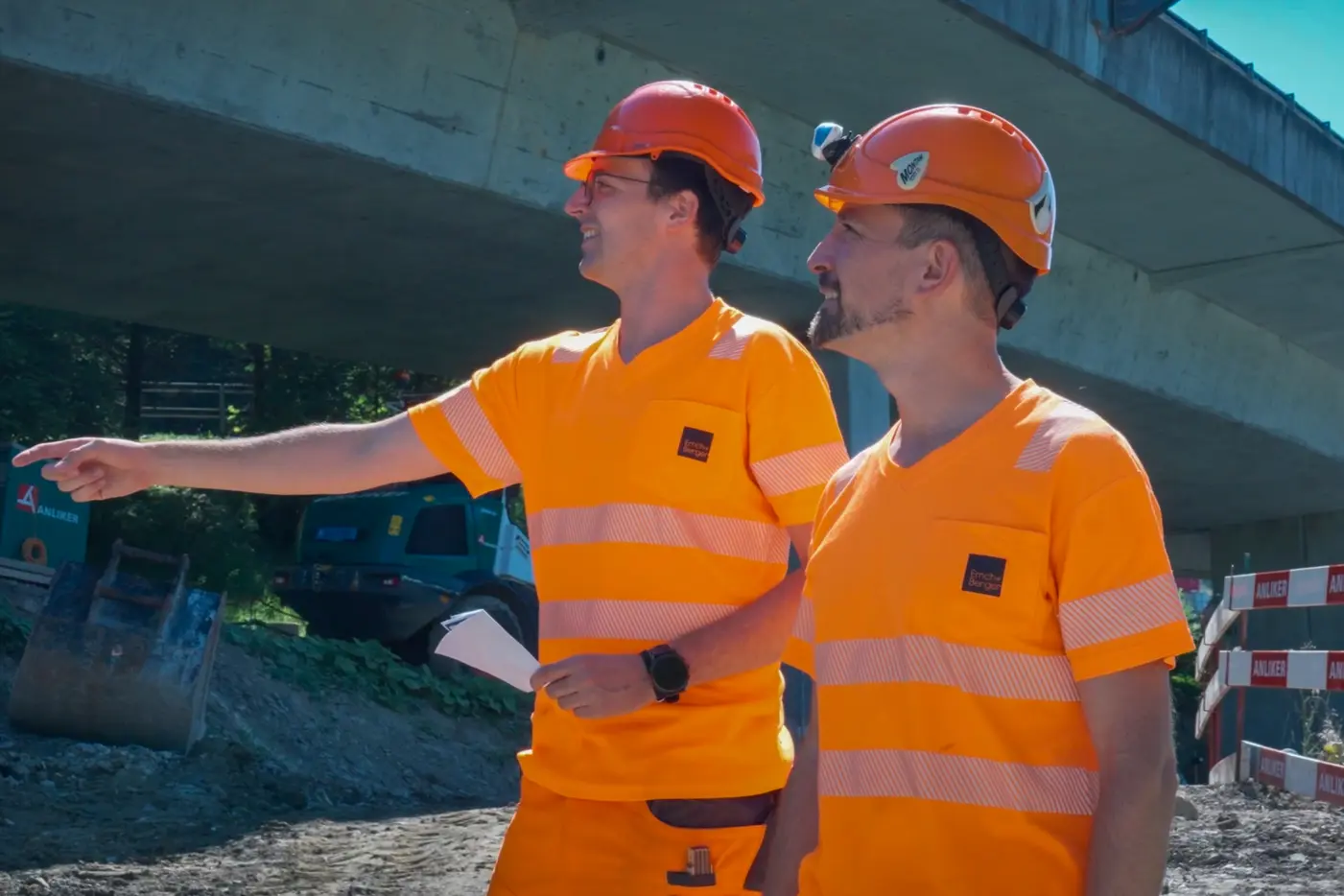 Zwei Bauarbeiter in leuchtend orangefarbener Arbeitskleidung stehen unter einer Brücke.