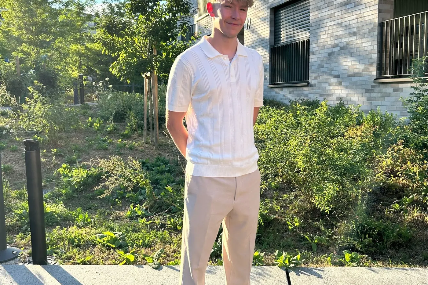 Junger Mann in einem weissen Poloshirt und beigen Hosen, vor einem modernen Gebäude.