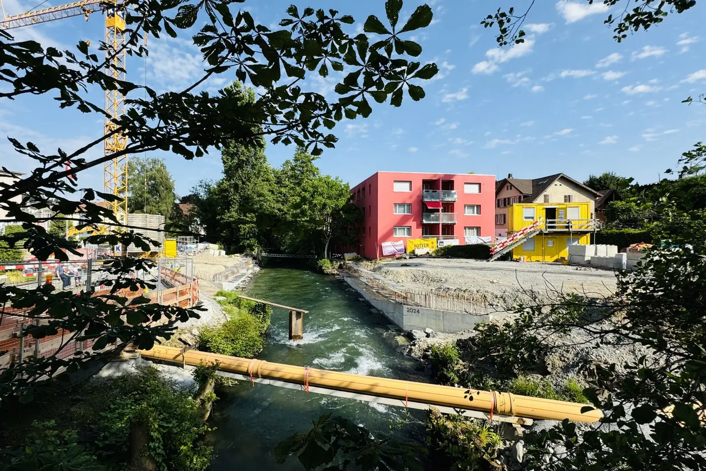 Blick auf einen Fluss mit mehreren Gebäuden und Bauarbeiten im Hintergrund unter blauem Himmel.