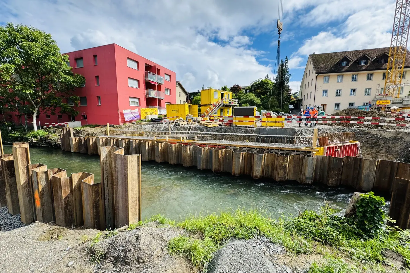 Baustelle am Fluss mit Holzplanken und bunten Containergebäuden, unter bewölktem Himmel.