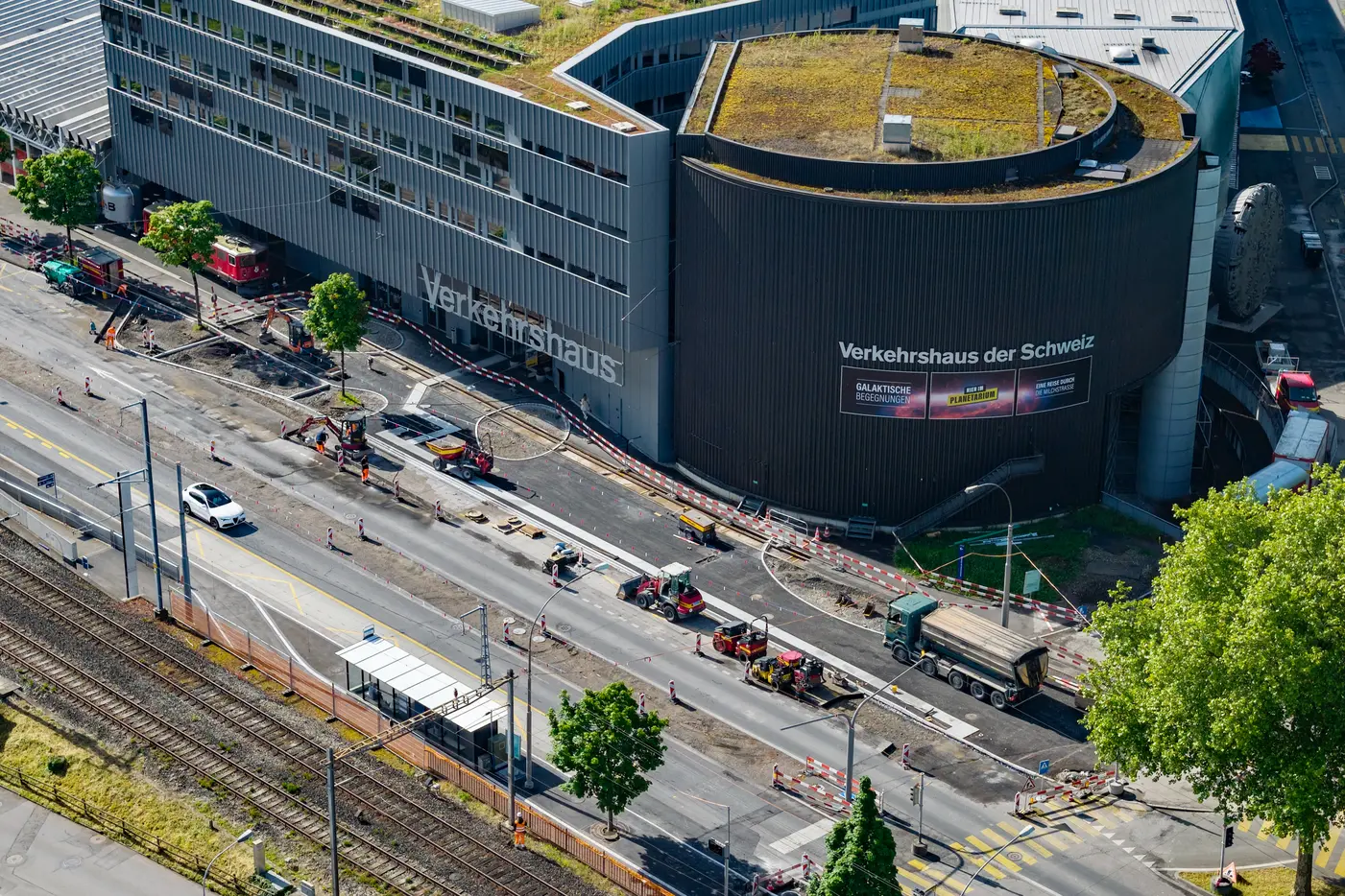 Blick auf eine Baustelle vor einem modernen Gebäude mit Straßenarbeiten und Fahrzeugen.