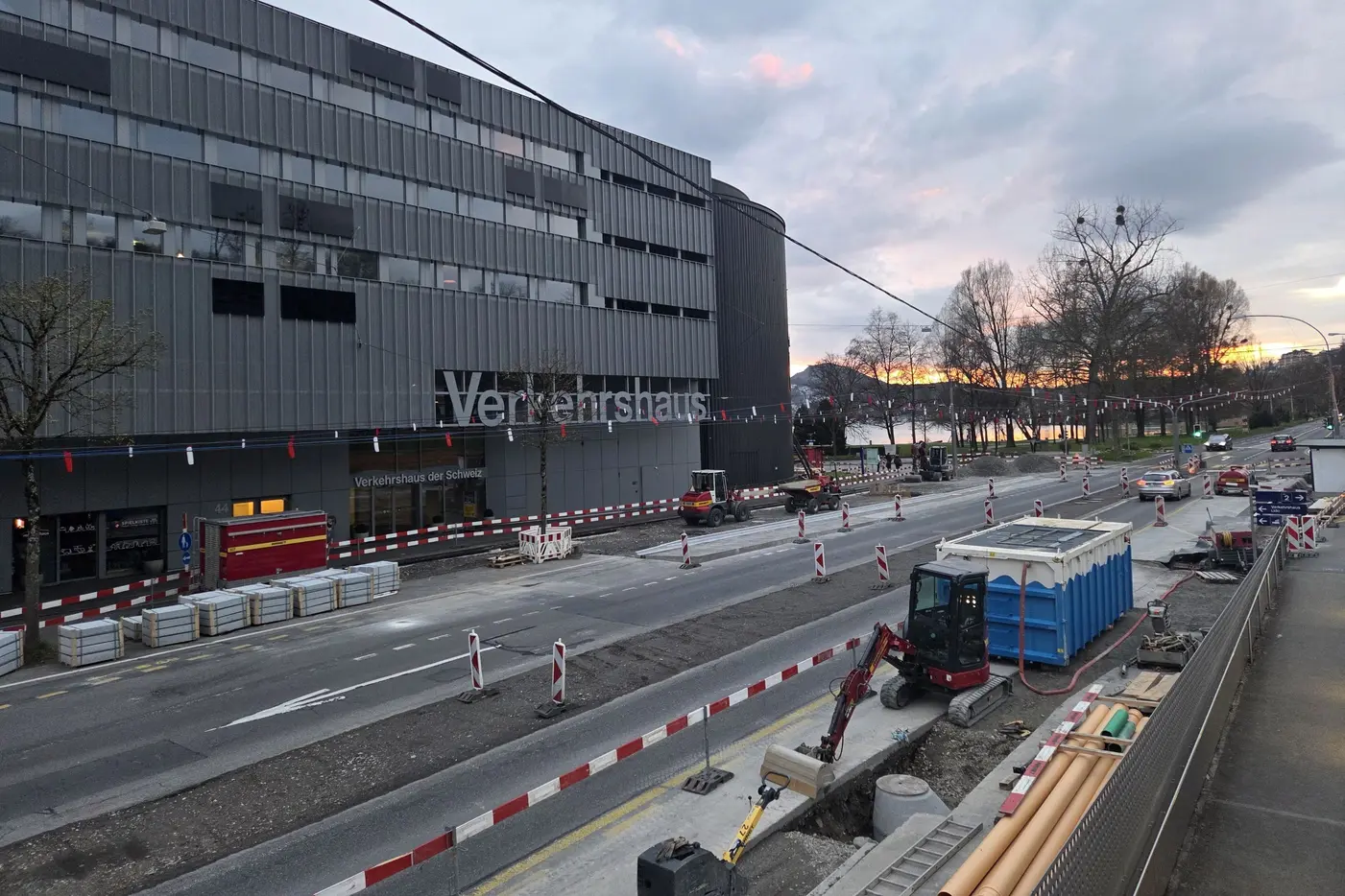 Bauarbeiten an einer Straße vor einem modernen Gebäude, Abendhimmel im Hintergrund.