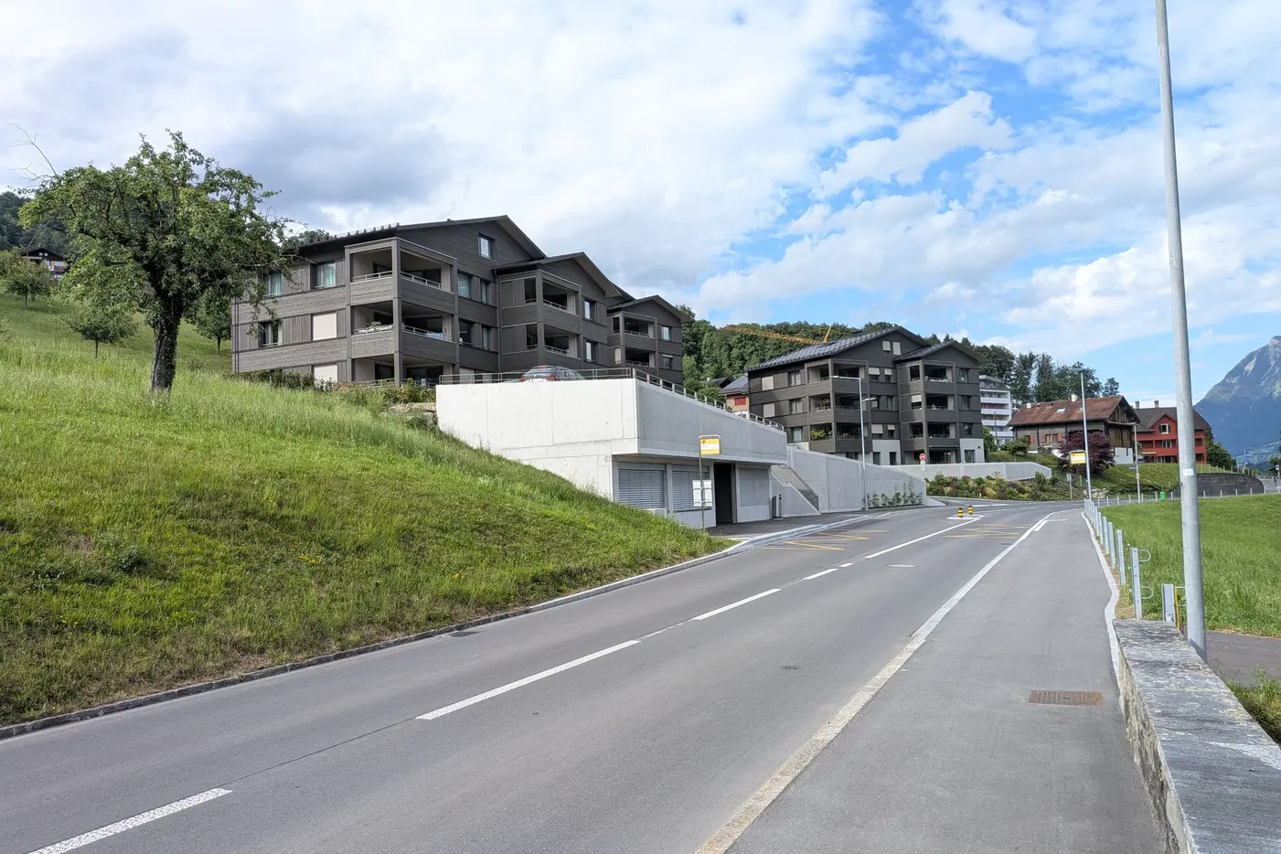 Moderne Wohngebäude an einer ruhigen Straße, umgeben von grünen Hügeln und blauer Himmel.