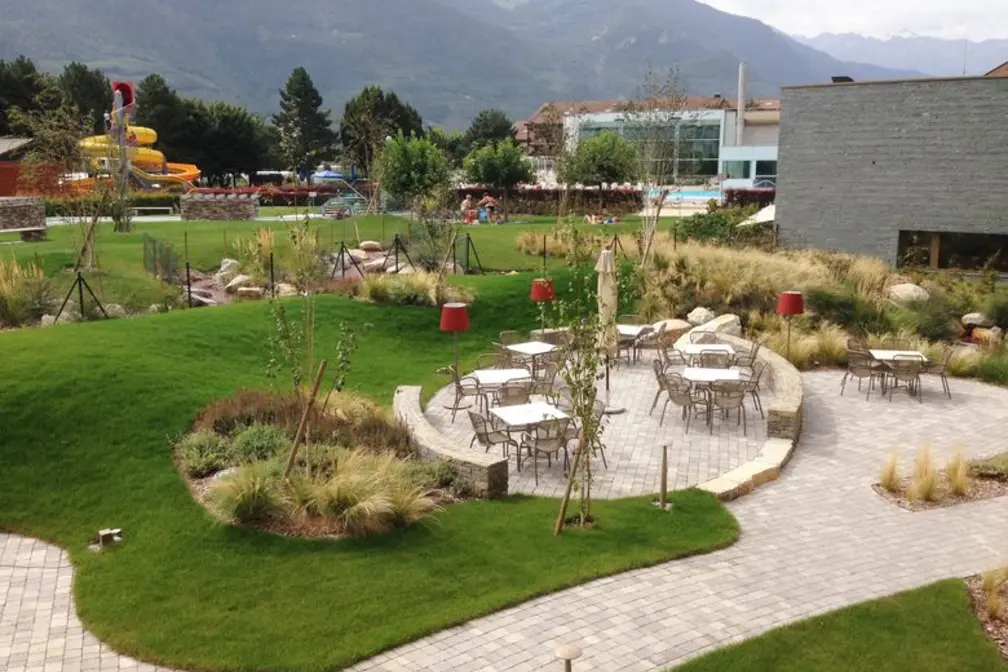 Grüne Gartenlandschaft mit Sitzplätzen, Palisaden und Bergen im Hintergrund.
