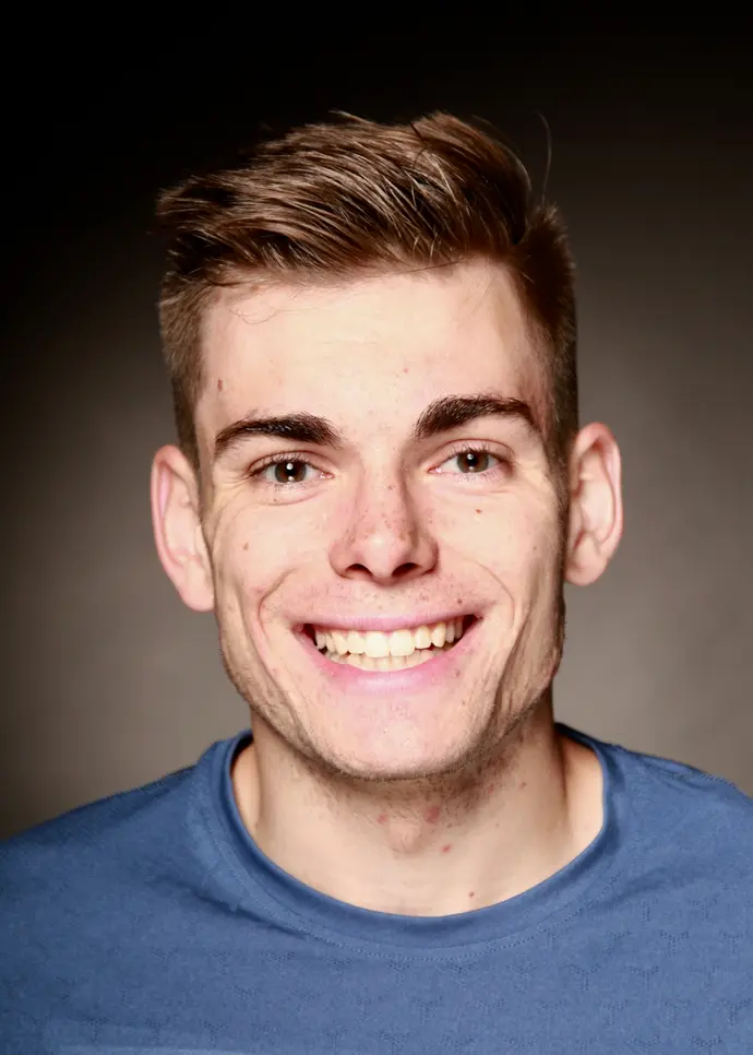Smiling young man with short hair dressed in a blue shirt against a dark background.