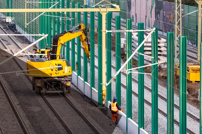 Baumaschinen und Arbeiter installieren grüne Stützpfeiler neben den Gleisen.